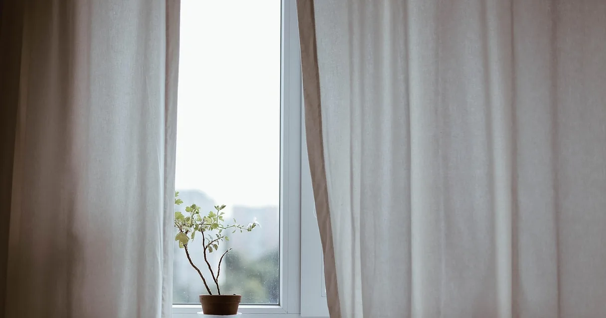 Cortinas claras ao lado de uma janela com um vasinho de planta, ilustrando como medir cortina para janela