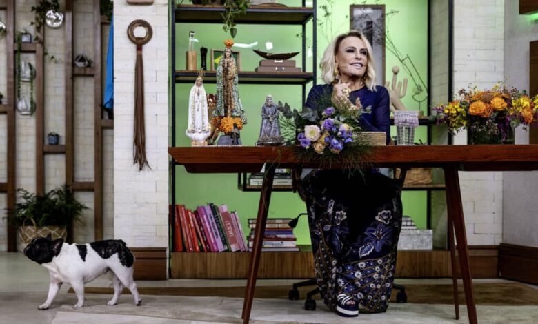 Uma apresentadora está sentada em uma mesa de madeira em um estúdio decorado. Ela usa um vestido azul escuro com detalhes em dourado e está cercada por arranjos de flores. Ao fundo, há prateleiras com livros e objetos decorativos. Um cão de pequeno porte, com pelagem branca e manchas pretas, caminha pelo chão ao lado da mesa.