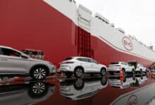 A imagem mostra um navio de carga da marca BYD, com a parte frontal vermelha e o logotipo visível. Na frente do navio, há vários carros estacionados, refletindo na água acumulada no chão. Os carros são de cor prata e possuem detalhes em azul. O ambiente parece ser um porto, com equipamentos e containers ao fundo.