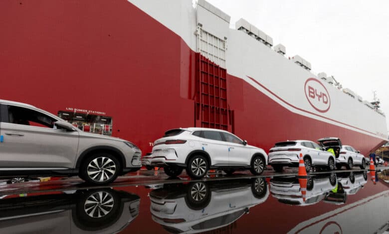 A imagem mostra um navio de carga da marca BYD, com a parte frontal vermelha e o logotipo visível. Na frente do navio, há vários carros estacionados, refletindo na água acumulada no chão. Os carros são de cor prata e possuem detalhes em azul. O ambiente parece ser um porto, com equipamentos e containers ao fundo.