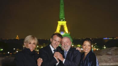 Quatro pessoas estão posando para a foto em frente à Torre Eiffel iluminada à noite. A torre está com luzes verdes e amarelas. As pessoas estão sorrindo e se abraçando, vestindo roupas de inverno. O fundo é uma vista noturna da cidade de Paris, com algumas luzes visíveis.