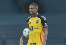 Um jogador de futebol está sorrindo enquanto segura uma bola. Ele veste uma camisa de goleiro predominantemente amarela com detalhes em preto e laranja, e o logotipo do time