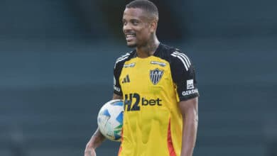 Um jogador de futebol está sorrindo enquanto segura uma bola. Ele veste uma camisa de goleiro predominantemente amarela com detalhes em preto e laranja, e o logotipo do time
