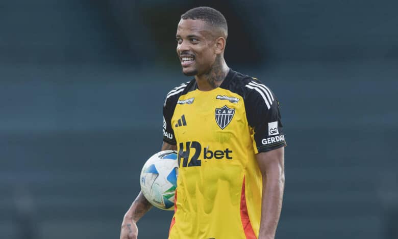 Um jogador de futebol está sorrindo enquanto segura uma bola. Ele veste uma camisa de goleiro predominantemente amarela com detalhes em preto e laranja, e o logotipo do time