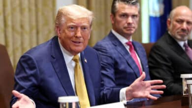 A imagem mostra um grupo de homens em uma sala de reuniões. Donald Trump está à esquerda, com um terno escuro e uma gravata amarela, gesticulando com as mãos. Ao centro, um homem com um terno azul claro observa, enquanto à direita, outro homem com um terno escuro e uma gravata rosa também está presente. A mesa está decorada com copos de água e há uma bandeira ao fundo.