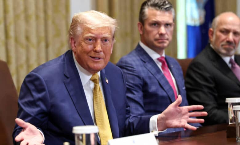 A imagem mostra um grupo de homens em uma sala de reuniões. Donald Trump está à esquerda, com um terno escuro e uma gravata amarela, gesticulando com as mãos. Ao centro, um homem com um terno azul claro observa, enquanto à direita, outro homem com um terno escuro e uma gravata rosa também está presente. A mesa está decorada com copos de água e há uma bandeira ao fundo.