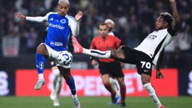 A imagem mostra um momento de um jogo de futebol, onde um jogador com uniforme azul e branco está tentando driblar um adversário que está em uma posição de chute. O jogador adversário, vestido com um uniforme preto e branco, está levantando a perna para interceptar a bola. Ao fundo, é possível ver um árbitro e outros jogadores, além de uma arquibancada com torcedores.