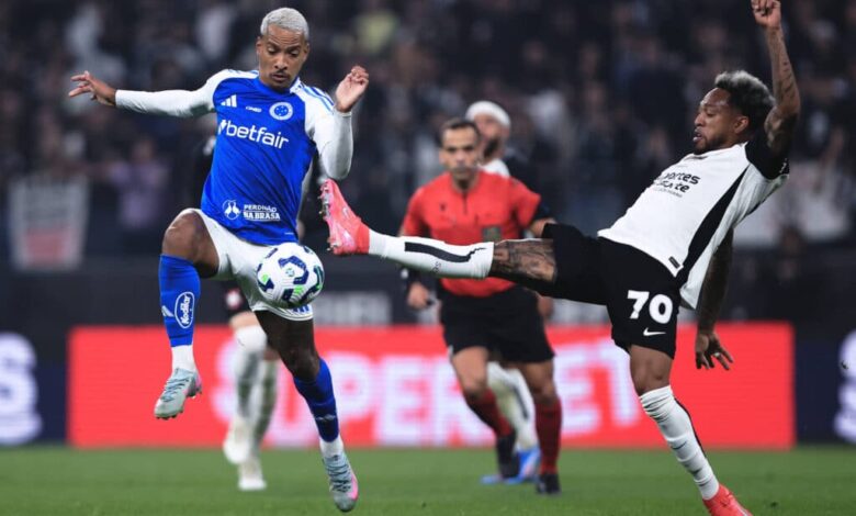 A imagem mostra um momento de um jogo de futebol, onde um jogador com uniforme azul e branco está tentando driblar um adversário que está em uma posição de chute. O jogador adversário, vestido com um uniforme preto e branco, está levantando a perna para interceptar a bola. Ao fundo, é possível ver um árbitro e outros jogadores, além de uma arquibancada com torcedores.