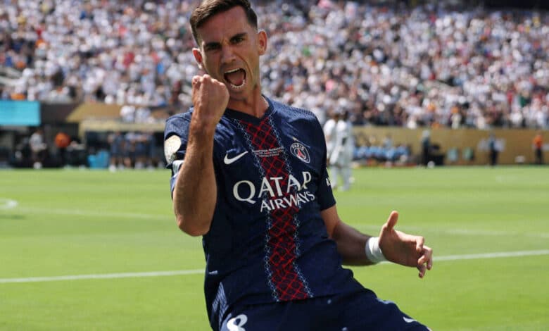 Um jogador de futebol está comemorando um gol em um campo. Ele veste um uniforme azul escuro com detalhes vermelhos e o logotipo da Qatar Airways. O jogador está com o punho cerrado e uma expressão de alegria. Ao fundo, há uma multidão de torcedores nas arquibancadas, com algumas pessoas vestindo camisetas brancas. O campo está bem iluminado, indicando que é um dia ensolarado.