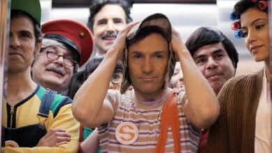 Um grupo de pessoas está dentro de um elevador. A maioria delas está vestida com roupas coloridas e fantasias, sugerindo um tema de festa ou evento. Um homem no centro parece estar um pouco desconfortável, segurando o chapéu na cabeça. As outras pessoas ao redor estão sorrindo e se divertindo. O ambiente é um elevador moderno, com paredes metálicas e iluminação suave.