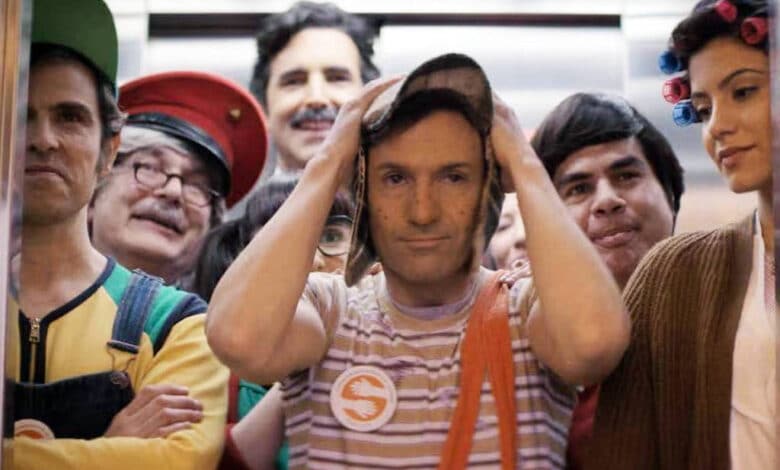 Um grupo de pessoas está dentro de um elevador. A maioria delas está vestida com roupas coloridas e fantasias, sugerindo um tema de festa ou evento. Um homem no centro parece estar um pouco desconfortável, segurando o chapéu na cabeça. As outras pessoas ao redor estão sorrindo e se divertindo. O ambiente é um elevador moderno, com paredes metálicas e iluminação suave.