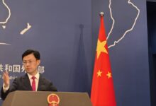Um homem em pé em um pódio, gesticulando com a mão. Ao fundo, uma bandeira da China com estrelas e um mapa do mundo desenhado na parede. O homem está vestido com um terno escuro e uma gravata vermelha. O pódio possui o emblema da República Popular da China.
