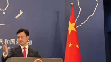 Um homem em pé em um pódio, gesticulando com a mão. Ao fundo, uma bandeira da China com estrelas e um mapa do mundo desenhado na parede. O homem está vestido com um terno escuro e uma gravata vermelha. O pódio possui o emblema da República Popular da China.