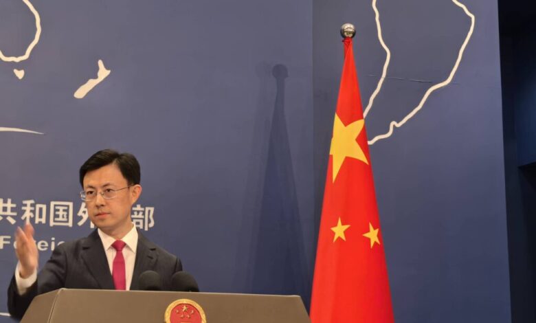 Um homem em pé em um pódio, gesticulando com a mão. Ao fundo, uma bandeira da China com estrelas e um mapa do mundo desenhado na parede. O homem está vestido com um terno escuro e uma gravata vermelha. O pódio possui o emblema da República Popular da China.