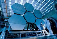 A couple of people wearing white outfits stand in front of the large mirrors of the James Webb Space Telescope
