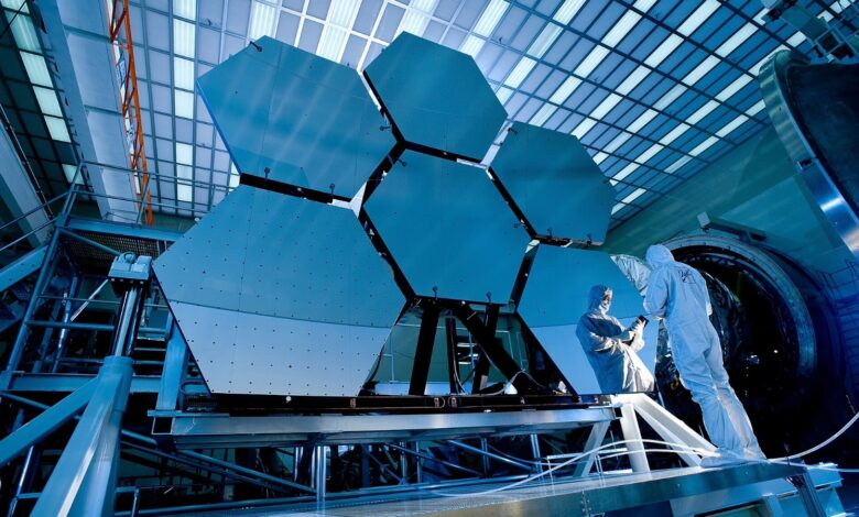 A couple of people wearing white outfits stand in front of the large mirrors of the James Webb Space Telescope