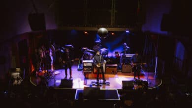 A imagem mostra uma banda se apresentando em um palco iluminado. Há vários músicos tocando instrumentos, incluindo guitarras e teclados. O palco é decorado com luzes coloridas e um grande disco de espelho pendurado no teto. A plateia é visível na parte inferior da imagem, com algumas pessoas assistindo ao show.