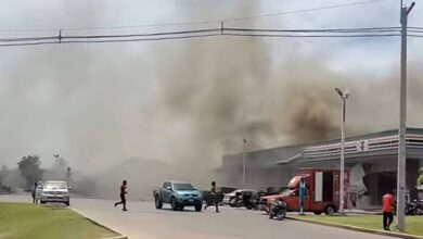 A imagem mostra uma cena de um incêndio em uma área urbana. Há uma grande quantidade de fumaça escura subindo no céu, proveniente de um edifício à direita. Algumas pessoas estão visíveis na rua, com um homem atravessando a via. Vários veículos estão estacionados e em movimento, incluindo um carro azul e um caminhão vermelho. O céu está parcialmente nublado e a vegetação é visível ao fundo.