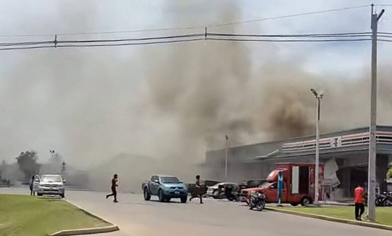 A imagem mostra uma cena de um incêndio em uma área urbana. Há uma grande quantidade de fumaça escura subindo no céu, proveniente de um edifício à direita. Algumas pessoas estão visíveis na rua, com um homem atravessando a via. Vários veículos estão estacionados e em movimento, incluindo um carro azul e um caminhão vermelho. O céu está parcialmente nublado e a vegetação é visível ao fundo.