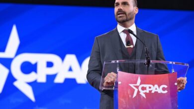 Um homem em um terno escuro está falando em um púlpito transparente com o logotipo do CPAC em destaque. O fundo é azul com o logotipo do CPAC em branco. O homem tem cabelo curto e barba bem aparada, e parece estar se dirigindo a uma audiência.