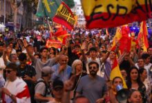 A imagem mostra uma grande multidão em uma manifestação, com pessoas levantando bandeiras da Espanha. Algumas pessoas estão sorrindo e acenando, enquanto outras seguram cartazes. O ambiente é festivo, com muitas pessoas vestindo camisetas e roupas em cores que representam a bandeira espanhola. Ao fundo, há edifícios e uma atmosfera de celebração.