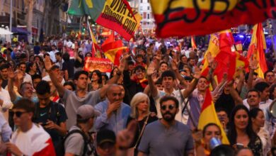 A imagem mostra uma grande multidão em uma manifestação, com pessoas levantando bandeiras da Espanha. Algumas pessoas estão sorrindo e acenando, enquanto outras seguram cartazes. O ambiente é festivo, com muitas pessoas vestindo camisetas e roupas em cores que representam a bandeira espanhola. Ao fundo, há edifícios e uma atmosfera de celebração.
