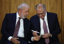 Na imagem, dois homens estão sentados lado a lado em um ambiente formal. O homem à esquerda tem cabelo grisalho e uma barba bem cuidada, vestindo um terno escuro com uma gravata listrada. O homem à direita, também com cabelo grisalho, usa óculos e um terno escuro com uma gravata roxa. Eles parecem estar conversando e o homem à direita está segurando um objeto em sua mão, enquanto o homem à esquerda observa atentamente.