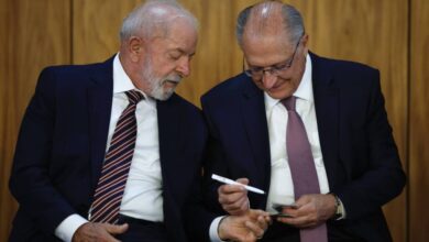 Na imagem, dois homens estão sentados lado a lado em um ambiente formal. O homem à esquerda tem cabelo grisalho e uma barba bem cuidada, vestindo um terno escuro com uma gravata listrada. O homem à direita, também com cabelo grisalho, usa óculos e um terno escuro com uma gravata roxa. Eles parecem estar conversando e o homem à direita está segurando um objeto em sua mão, enquanto o homem à esquerda observa atentamente.