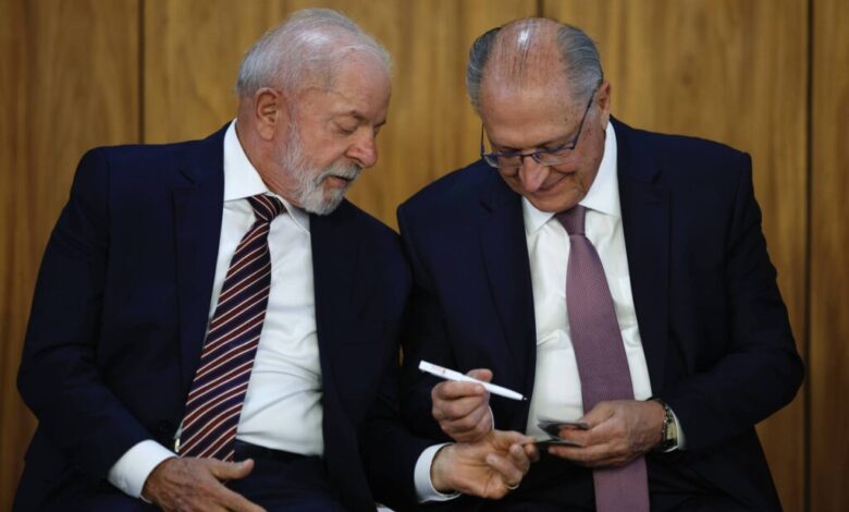 Na imagem, dois homens estão sentados lado a lado em um ambiente formal. O homem à esquerda tem cabelo grisalho e uma barba bem cuidada, vestindo um terno escuro com uma gravata listrada. O homem à direita, também com cabelo grisalho, usa óculos e um terno escuro com uma gravata roxa. Eles parecem estar conversando e o homem à direita está segurando um objeto em sua mão, enquanto o homem à esquerda observa atentamente.