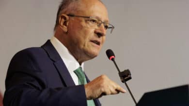 Um homem de óculos, com cabelo grisalho e calvo, está falando em um microfone. Ele usa um terno escuro com uma gravata verde e gesticula com a mão direita enquanto se expressa. O fundo é neutro e desfocado.