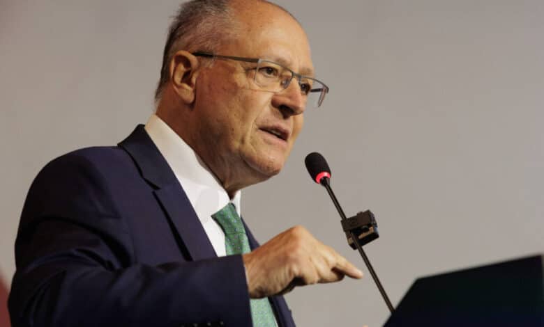 Um homem de óculos, com cabelo grisalho e calvo, está falando em um microfone. Ele usa um terno escuro com uma gravata verde e gesticula com a mão direita enquanto se expressa. O fundo é neutro e desfocado.