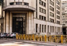 A imagem mostra a fachada de um edifício que abriga o INSS (Instituto Nacional do Seguro Social). O prédio é de estilo arquitetônico moderno, com uma entrada principal destacada por portas grandes e uma inscrição visível com as letras