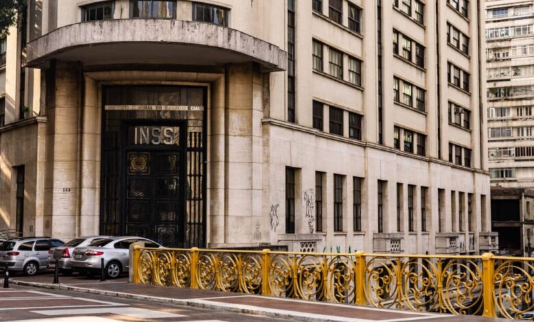 A imagem mostra a fachada de um edifício que abriga o INSS (Instituto Nacional do Seguro Social). O prédio é de estilo arquitetônico moderno, com uma entrada principal destacada por portas grandes e uma inscrição visível com as letras