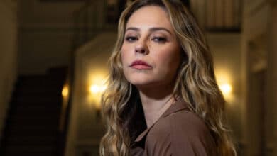 A imagem mostra uma mulher com cabelo longo e ondulado, usando uma blusa marrom. Ela está em um ambiente interno elegante, com uma escada ao fundo e um lustre iluminado. A mulher está em um ângulo ligeiramente de perfil, com uma expressão séria e as mãos unidas à frente do corpo.