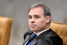 Um homem com cabelo curto e grisalho está sentado em uma cadeira de tribunal. Ele usa uma toga preta e uma gravata azul. O fundo é desfocado, mas parece ser um ambiente de tribunal, com cadeiras de couro marrom ao redor. O homem tem uma expressão séria e está olhando para o lado.