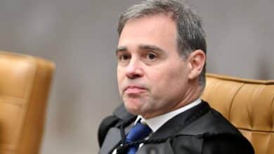 Um homem com cabelo curto e grisalho está sentado em uma cadeira de tribunal. Ele usa uma toga preta e uma gravata azul. O fundo é desfocado, mas parece ser um ambiente de tribunal, com cadeiras de couro marrom ao redor. O homem tem uma expressão séria e está olhando para o lado.