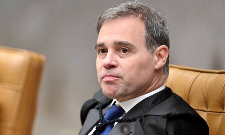 Um homem com cabelo curto e grisalho está sentado em uma cadeira de tribunal. Ele usa uma toga preta e uma gravata azul. O fundo é desfocado, mas parece ser um ambiente de tribunal, com cadeiras de couro marrom ao redor. O homem tem uma expressão séria e está olhando para o lado.