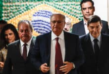 A imagem mostra um grupo de homens e mulheres em um ambiente formal. No centro, um homem de terno escuro e gravata vermelha caminha em direção à câmera, enquanto outros dois homens, um à sua esquerda e outro à direita, também estão vestidos formalmente. Ao fundo, há uma parede decorada com um padrão que remete à bandeira do Brasil, e algumas pessoas estão visíveis atrás deles. O ambiente parece ser um espaço de reunião ou evento oficial.