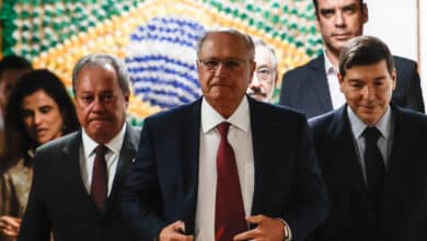 A imagem mostra um grupo de homens e mulheres em um ambiente formal. No centro, um homem de terno escuro e gravata vermelha caminha em direção à câmera, enquanto outros dois homens, um à sua esquerda e outro à direita, também estão vestidos formalmente. Ao fundo, há uma parede decorada com um padrão que remete à bandeira do Brasil, e algumas pessoas estão visíveis atrás deles. O ambiente parece ser um espaço de reunião ou evento oficial.