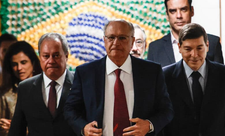 A imagem mostra um grupo de homens e mulheres em um ambiente formal. No centro, um homem de terno escuro e gravata vermelha caminha em direção à câmera, enquanto outros dois homens, um à sua esquerda e outro à direita, também estão vestidos formalmente. Ao fundo, há uma parede decorada com um padrão que remete à bandeira do Brasil, e algumas pessoas estão visíveis atrás deles. O ambiente parece ser um espaço de reunião ou evento oficial.