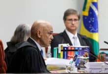 A imagem mostra uma sessão judicial com três pessoas. À esquerda, um homem calvo, vestido com uma toga preta e gravata, está falando. Ao centro, uma mulher com cabelo grisalho, também vestida com toga, escuta atentamente. À direita, um homem com cabelo curto e óculos, vestido com uma toga preta e camisa branca, observa. Ao fundo, há uma bandeira do Brasil e várias pastas de documentos sobre a mesa.