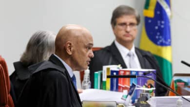 A imagem mostra uma sessão judicial com três pessoas. À esquerda, um homem calvo, vestido com uma toga preta e gravata, está falando. Ao centro, uma mulher com cabelo grisalho, também vestida com toga, escuta atentamente. À direita, um homem com cabelo curto e óculos, vestido com uma toga preta e camisa branca, observa. Ao fundo, há uma bandeira do Brasil e várias pastas de documentos sobre a mesa.