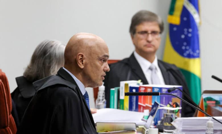 A imagem mostra uma sessão judicial com três pessoas. À esquerda, um homem calvo, vestido com uma toga preta e gravata, está falando. Ao centro, uma mulher com cabelo grisalho, também vestida com toga, escuta atentamente. À direita, um homem com cabelo curto e óculos, vestido com uma toga preta e camisa branca, observa. Ao fundo, há uma bandeira do Brasil e várias pastas de documentos sobre a mesa.