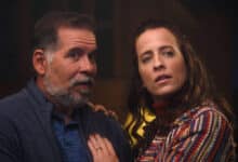 A imagem mostra um homem e uma mulher em uma cena que parece ser de tensão ou drama. O homem, com cabelo grisalho e barba, está usando uma camisa azul e um casaco escuro. Ele parece surpreso ou preocupado. A mulher, com cabelo castanho e ondulado, está vestindo uma blusa colorida e também parece estar em estado de alerta. Ambos estão olhando para algo fora do quadro, sugerindo que estão reagindo a uma situação intensa.