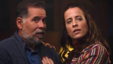 A imagem mostra um homem e uma mulher em uma cena que parece ser de tensão ou drama. O homem, com cabelo grisalho e barba, está usando uma camisa azul e um casaco escuro. Ele parece surpreso ou preocupado. A mulher, com cabelo castanho e ondulado, está vestindo uma blusa colorida e também parece estar em estado de alerta. Ambos estão olhando para algo fora do quadro, sugerindo que estão reagindo a uma situação intensa.
