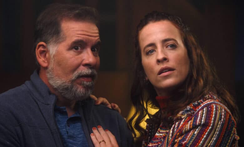 A imagem mostra um homem e uma mulher em uma cena que parece ser de tensão ou drama. O homem, com cabelo grisalho e barba, está usando uma camisa azul e um casaco escuro. Ele parece surpreso ou preocupado. A mulher, com cabelo castanho e ondulado, está vestindo uma blusa colorida e também parece estar em estado de alerta. Ambos estão olhando para algo fora do quadro, sugerindo que estão reagindo a uma situação intensa.