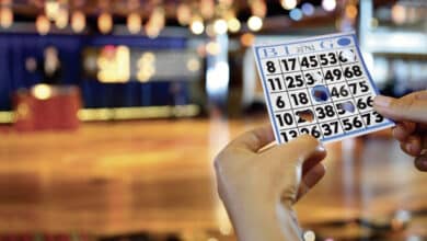 A imagem mostra uma mão segurando uma cartela de bingo com números dispostos em colunas marcadas com as letras B, I, N, G e O. Ao fundo, há um ambiente de jogo com mesas e luzes decorativas, sugerindo um evento de bingo. O chão é de madeira e há um palco ao fundo com um apresentador.