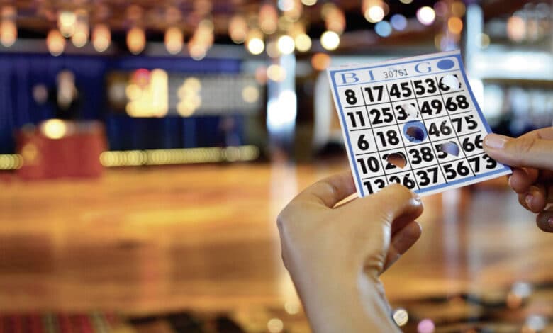 A imagem mostra uma mão segurando uma cartela de bingo com números dispostos em colunas marcadas com as letras B, I, N, G e O. Ao fundo, há um ambiente de jogo com mesas e luzes decorativas, sugerindo um evento de bingo. O chão é de madeira e há um palco ao fundo com um apresentador.