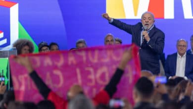 A imagem mostra um evento político com uma multidão. Um homem de cabelo grisalho e barba, vestido com um terno escuro, está falando ao microfone. Ele gesticula com a mão direita. Ao fundo, há pessoas sentadas, algumas delas olhando para o orador. Na frente, uma faixa rosa é visível, com texto em letras brancas, mas o conteúdo do texto não é legível. O fundo é colorido, com predominância de azul e amarelo.