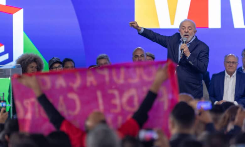 A imagem mostra um evento político com uma multidão. Um homem de cabelo grisalho e barba, vestido com um terno escuro, está falando ao microfone. Ele gesticula com a mão direita. Ao fundo, há pessoas sentadas, algumas delas olhando para o orador. Na frente, uma faixa rosa é visível, com texto em letras brancas, mas o conteúdo do texto não é legível. O fundo é colorido, com predominância de azul e amarelo.